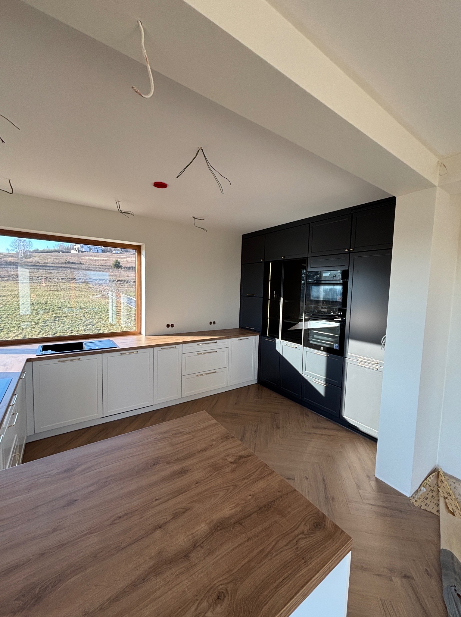 Nowoczesna kuchnia z białymi szafkami dolnymi i czarnymi górnymi, drewniany blat i podłoga w jodełkę. Widoczne niedokończone instalacje elektryczne.