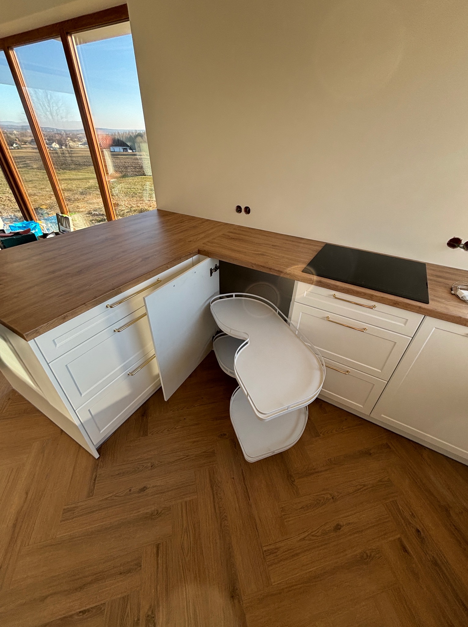 Biała kuchnia z drewnianym blatem i wysuwaną półką narożną, widok z góry na parkiet w jodełkę. Duże okno z widokiem na krajobraz.