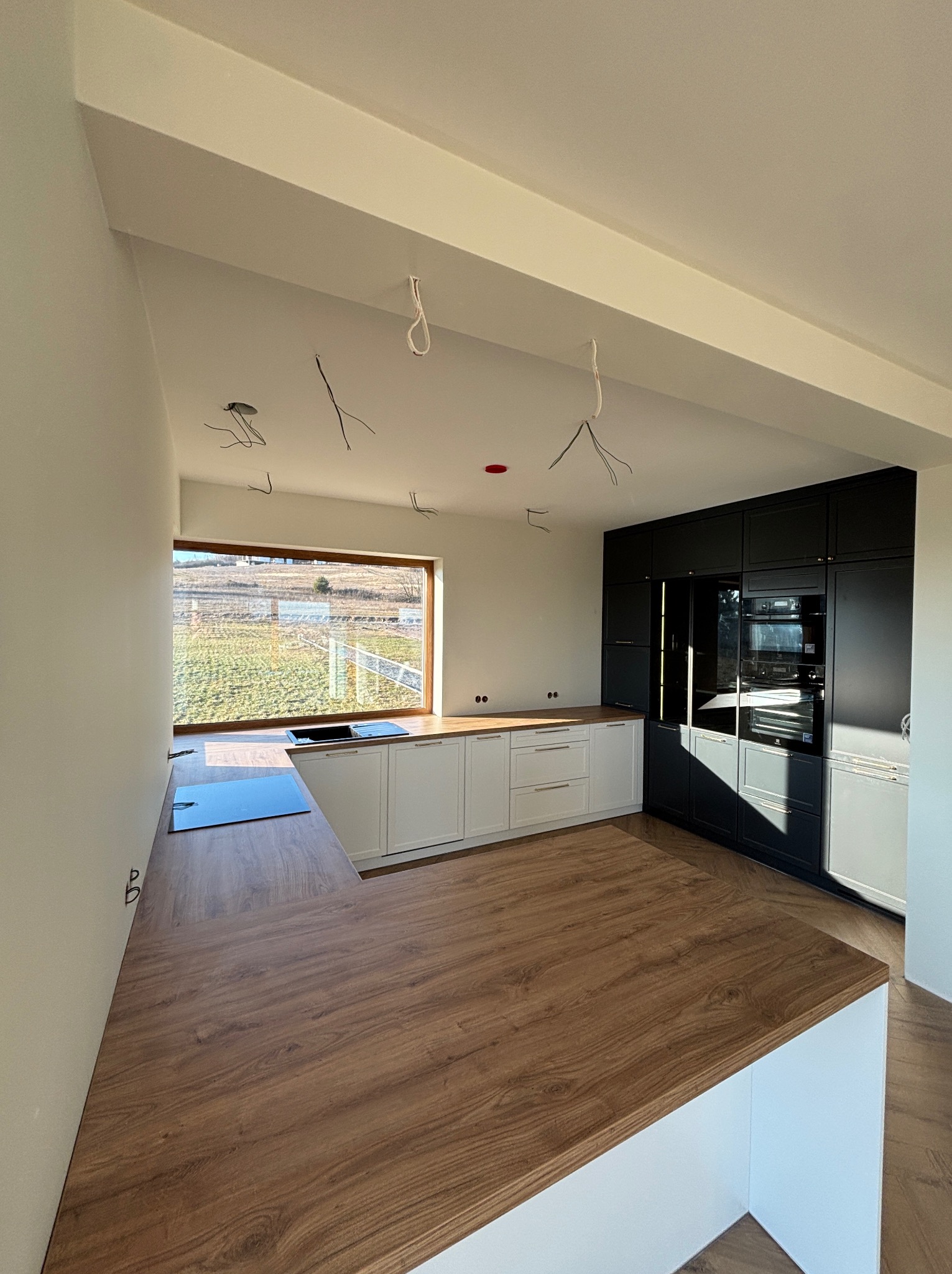 Nowoczesna kuchnia z białymi szafkami dolnymi i czarnymi górnymi, drewniany blat, duże okno z widokiem na pole. Wnętrze w trakcie wykończenia, widoczne kable.