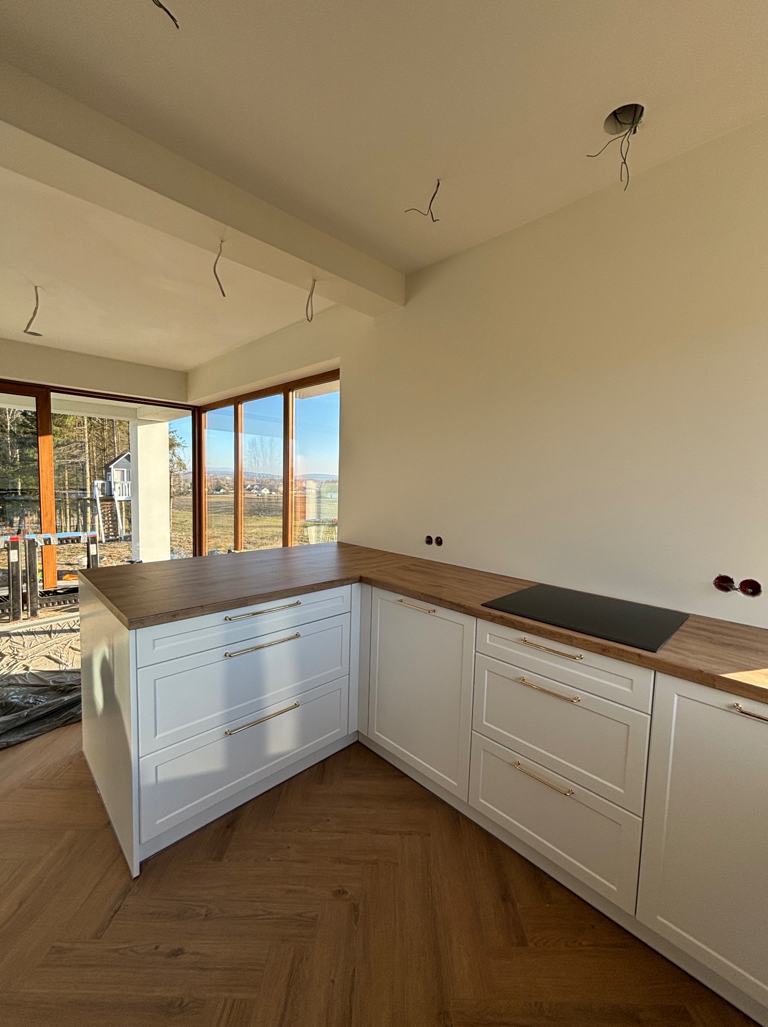 Biała zabudowa kuchenna z drewnianym blatem i złotymi uchwytami, w surowym stanie, z widokiem na zewnątrz w Kielcach. Ułożona podłoga w jodełkę.