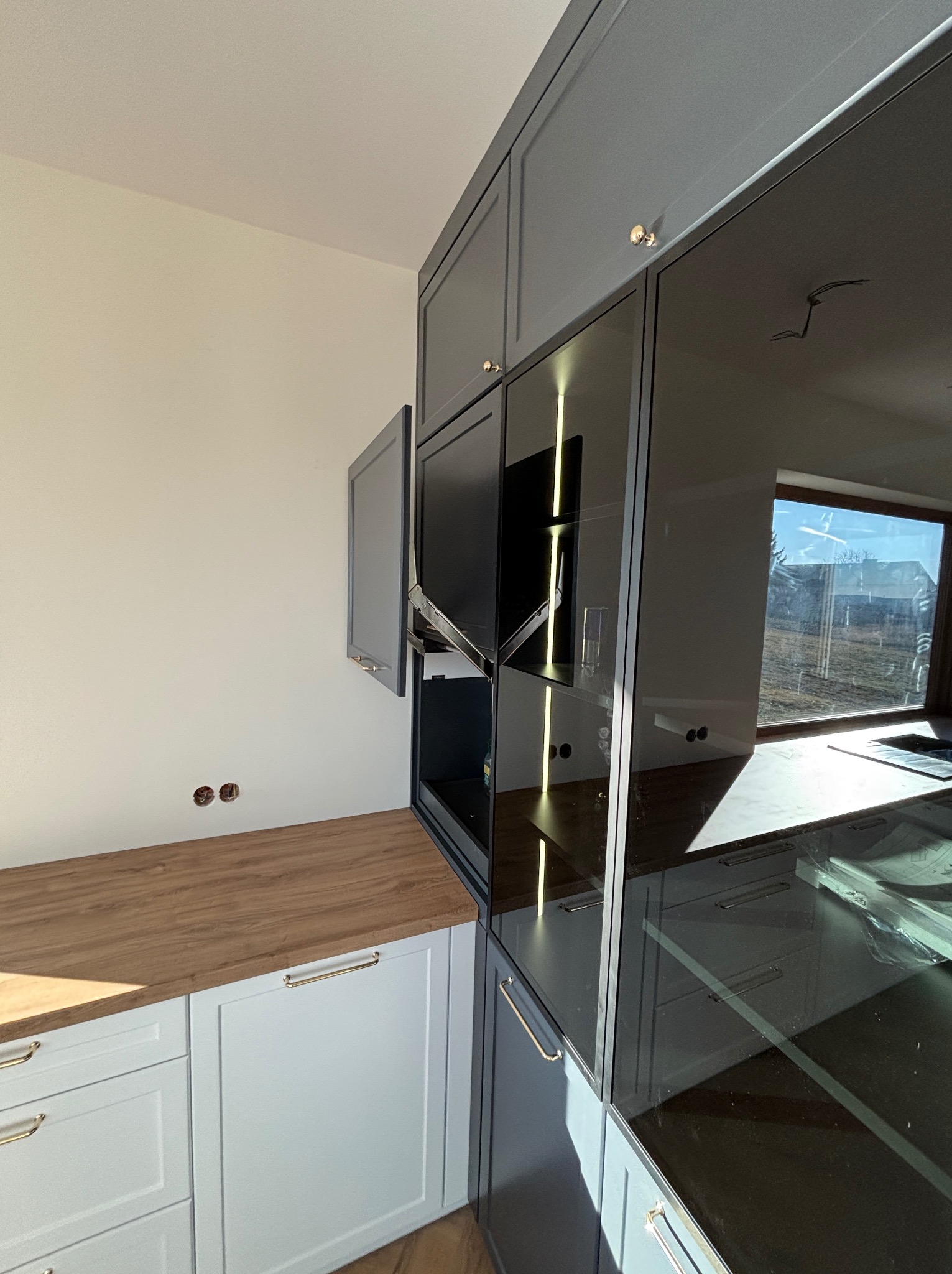 Szara zabudowa kuchenna z drewnianym blatem i szklanymi frontami szafek, widoczne otwory na kable w ścianie, złote uchwyty, nowoczesny design.