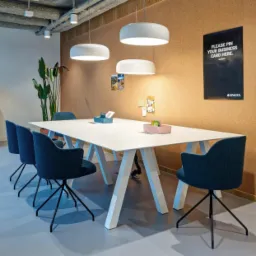 Nowoczesna sala konferencyjna z dużym białym stołem na trójkątnych nogach, otoczonym granatowymi krzesłami na czarnych nogach, z korkową ścianą, wiszącymi lampami i rośliną doniczkową w tle.