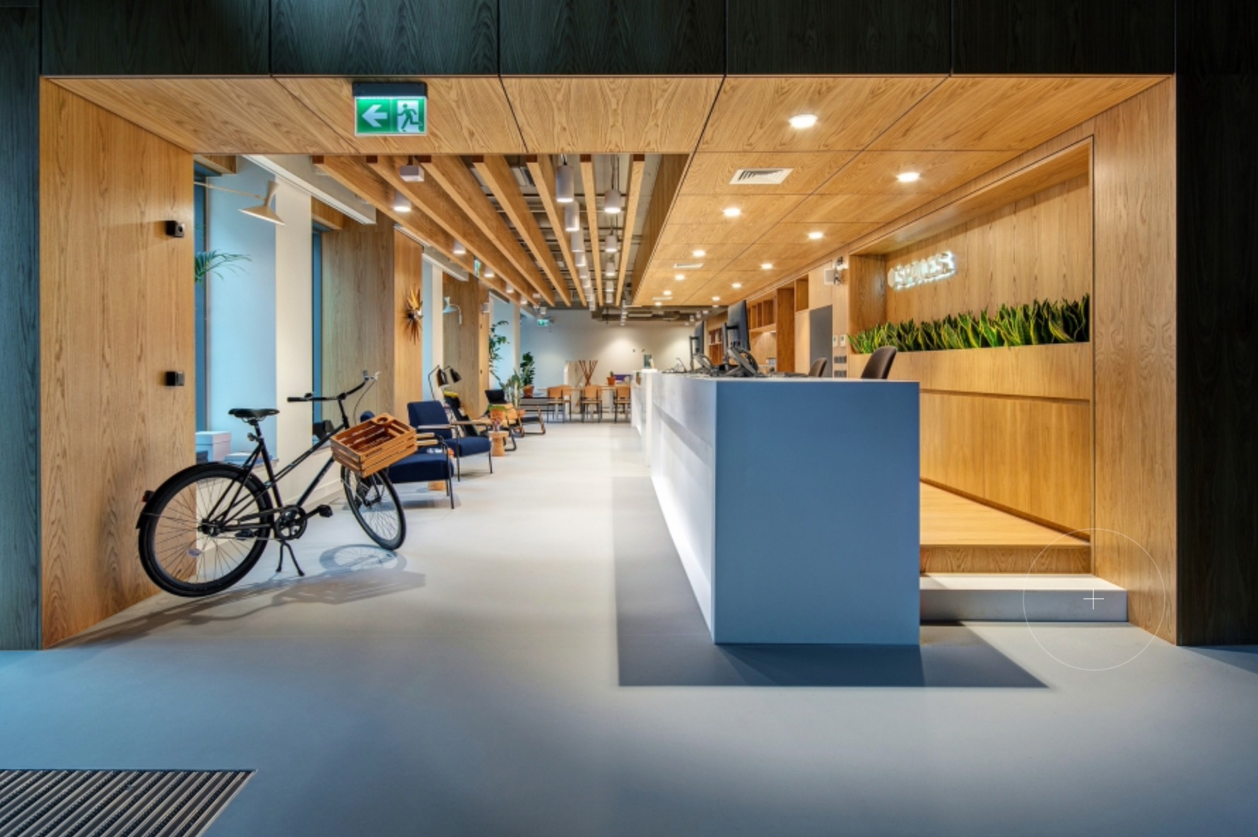 Nowoczesne biuro z drewnianymi elementami, rower z koszykiem przy wejściu, recepcja z białym blatem, oświetlenie sufitowe z drewnianymi belkami, rośliny doniczkowe i krzesła w tle.