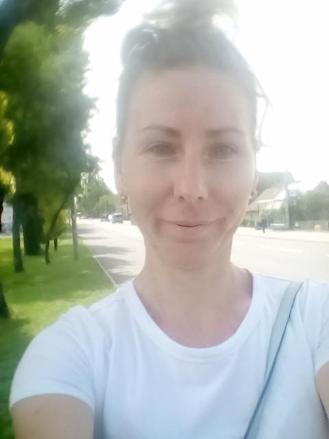 Portret kobiety w białej koszulce, z rozmazanym tłem ulicy i zieleni, sfotografowany w jasnym świetle słonecznym.