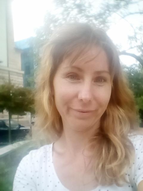 Portret kobiety z blond włosami w koszulce w kropki, na tle budynku i zieleni. Zdjęcie wykonane z bliska, w naturalnym oświetleniu.