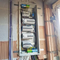 Rozdzielnica elektryczna z zabezpieczeniami zamontowana na ścianie z częściowo wykończonym murem, widoczne kable prowadzone w korytach.