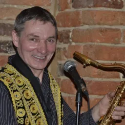 Uśmiechnięty muzyk z saksofonem i mikrofonem na tle ceglanej ściany, ubrany w czarną koszulę w paski i ozdobną kamizelkę z motywami geometrycznymi w kolorze złota.