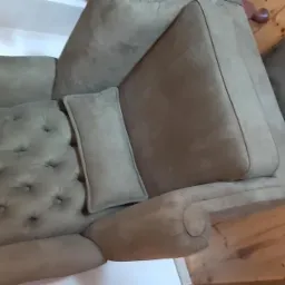 Odnowiony fotel z zieloną tapicerką, pikowany na oparciu, z poduszką lędźwiową, stojący na drewnianej podłodze.