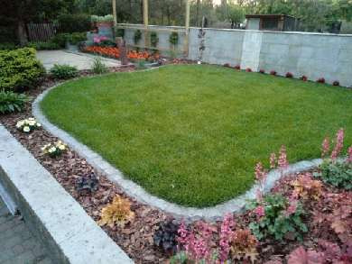 Kwadratowy trawnik otoczony ozdobnym obrzeżem z betonu i kory, z rabatami kwiatowymi wzdłuż ścieżki i muru w tle.