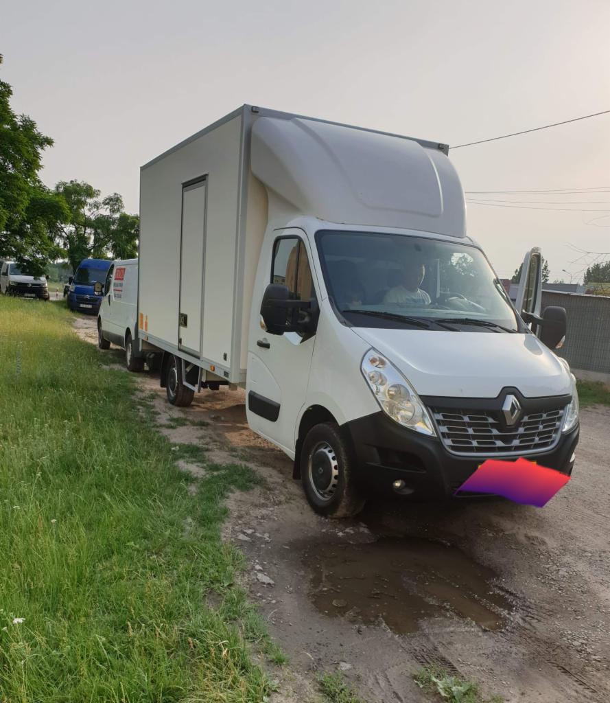 Biały samochód dostawczy Renault Master z otwartymi drzwiami kierowcy, stojący na polnej drodze z kałużą, w tle zaparkowane inne busy.
