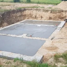 Wylane betonowe fundamenty domu jednorodzinnego w wykopie, widoczne pomarańczowe rurki pionowe, na ziemi leży kielnia, w tle stosy ziemi.