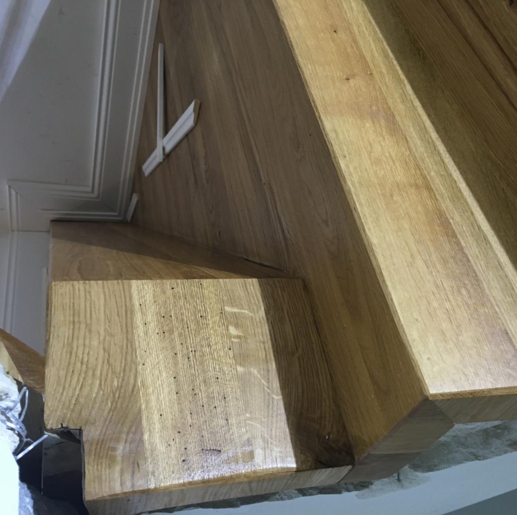 Szczegółowe ujęcie drewnianych schodów z widocznymi drobnymi otworami po szkodnikach w drewnie, fragment listwy przypodłogowej i ściana w tle.