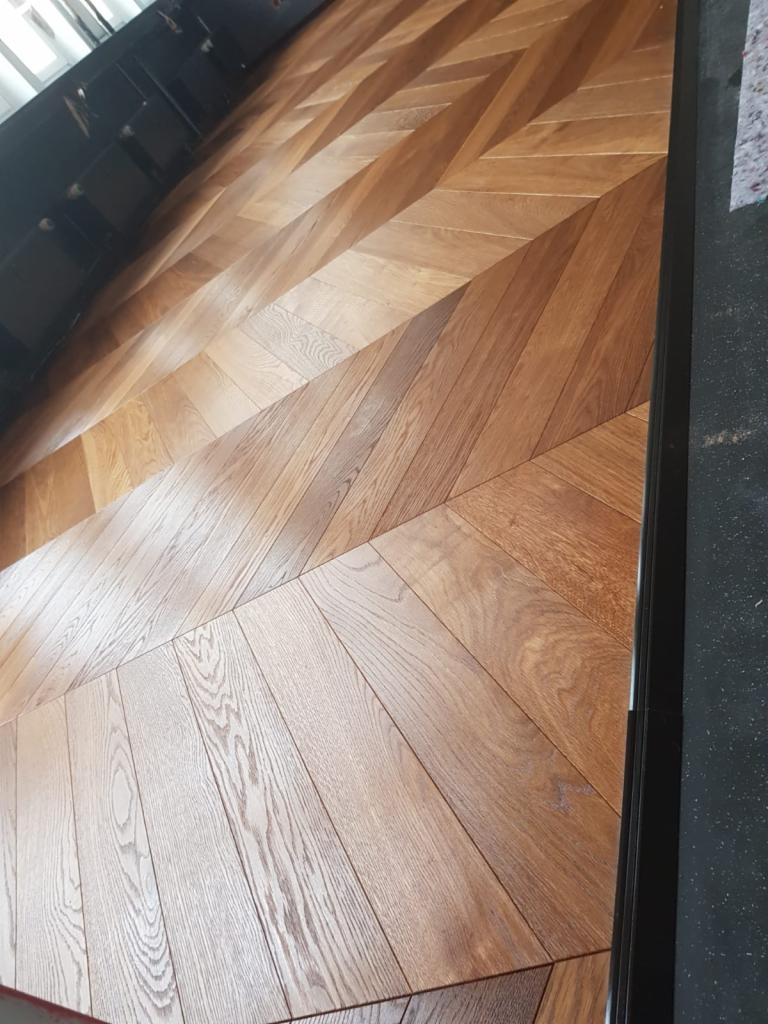 Ułożony parkiet w jodełkę, wykonany z drewna o zróżnicowanym odcieniu, z widoczną ramą wykończeniową i fragmentem czarnej podłogi w tle.