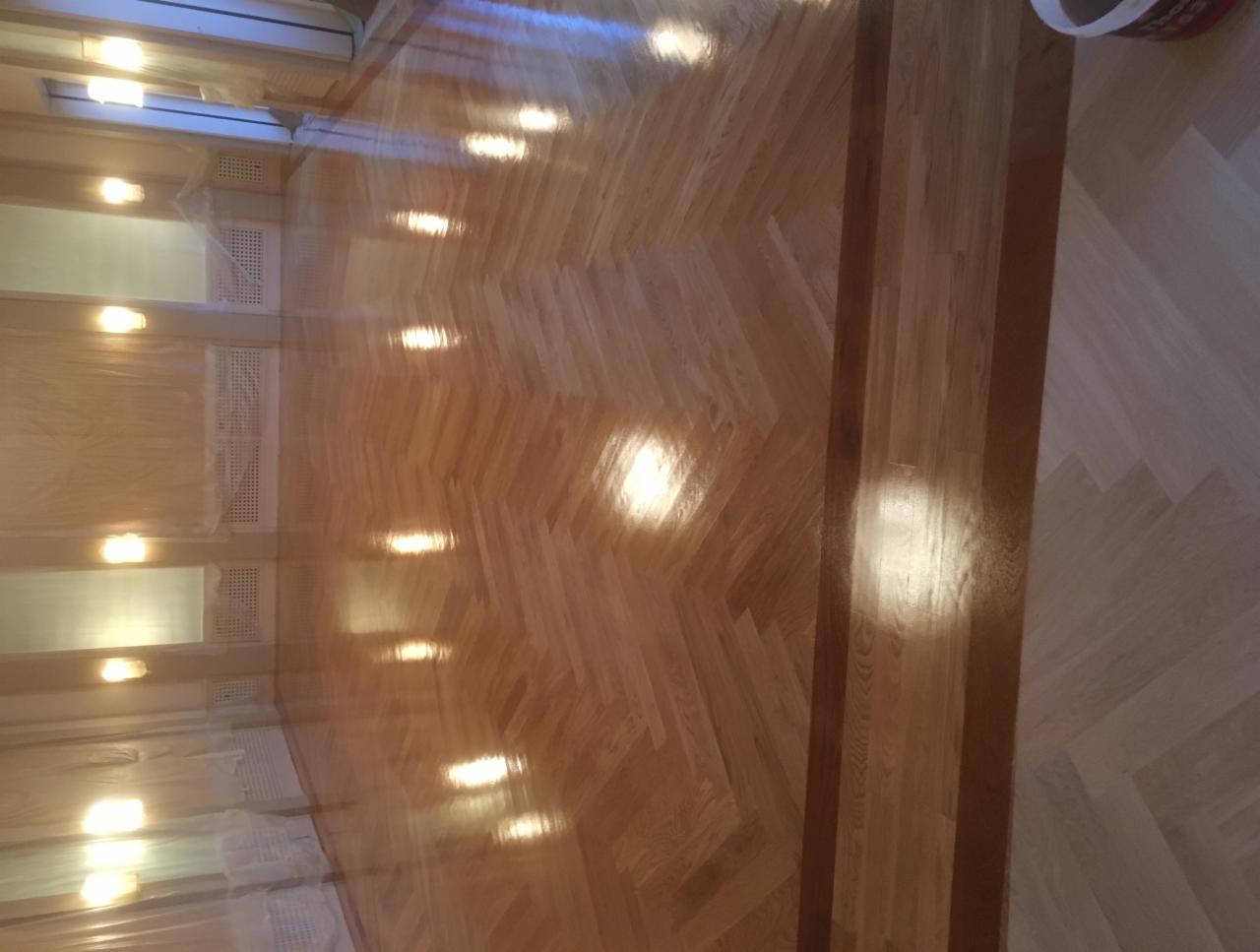 Błyszcząca, lakierowana podłoga parkietowa ułożona we wzór jodełki z ciemniejszą obwódką, okna zabezpieczone folią malarską.