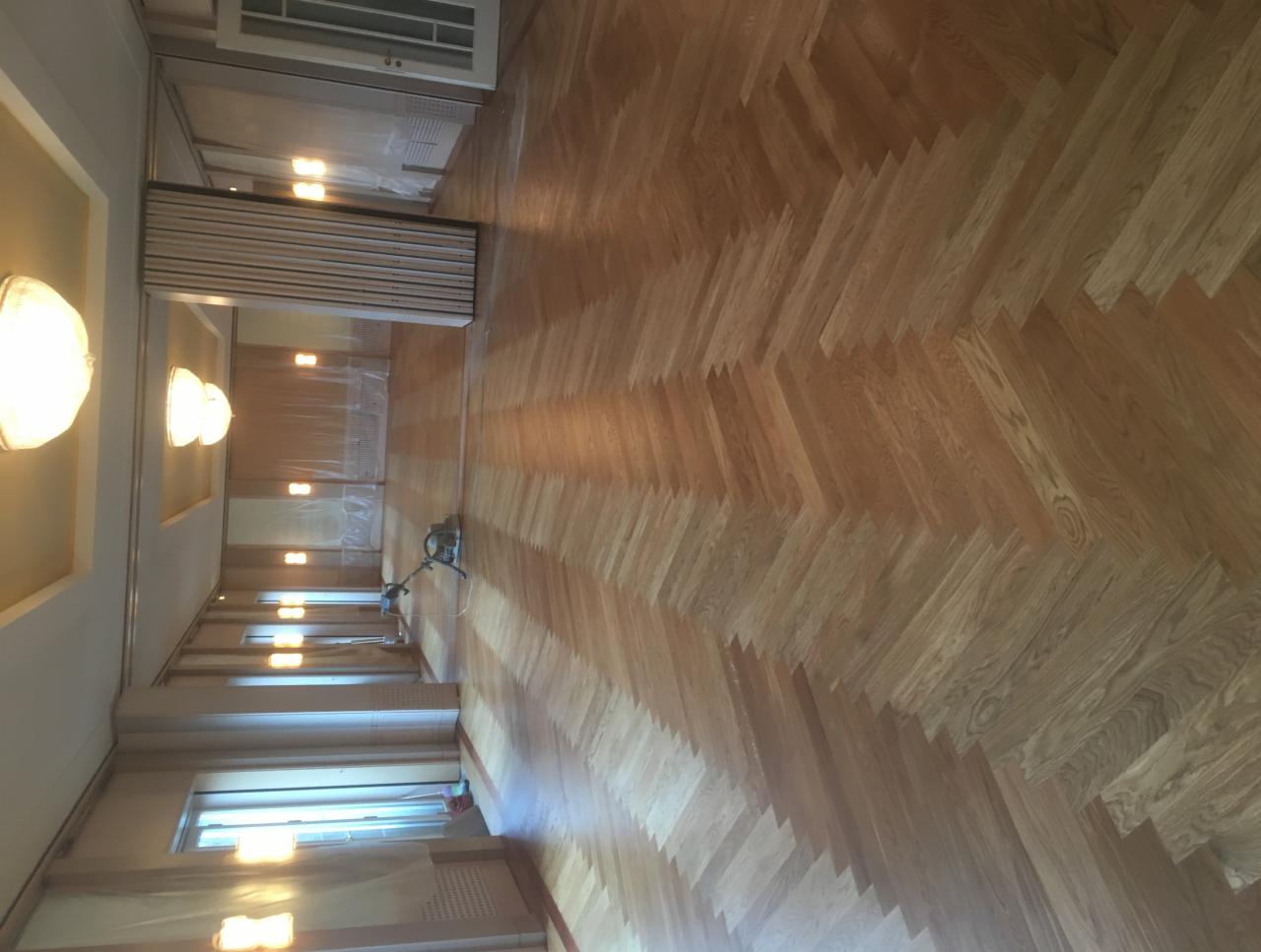 Parkiet ułożony w jodełkę, częściowo wycyklinowany, z widoczną maszyną do cyklinowania w tle, oświetlony ciepłym światłem lamp sufitowych.