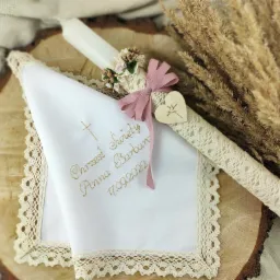 Biała, haftowana chusteczka z imieniem i datą chrztu obok ozdobnej świecy z kokardą i drewnianym sercem, ułożone na drewnianym plastrze, z dekoracją z suszonych traw w tle.