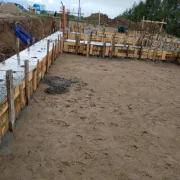 Wykop pod fundamenty z deskowaniem i zalanym betonem. Widoczne wystające pręty zbrojeniowe, rury instalacyjne i ślady stóp na piasku. Prace budowlane w toku.