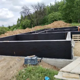 Zabezpieczone, czarne fundamenty budynku w trakcie budowy. Widoczne zbrojenia, izolacja pionowa i teren wokół fundamentów. Na pierwszym planie ubijarka.