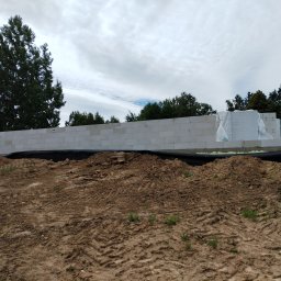 Baldyg Bud - Wznoszone ściany z białych bloczków na fundamencie, zabezpieczone czarną folią. Na paletach widoczne materiały budowlane w folii ochronnej. Krajobraz z drzewami w tle.