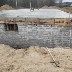 Baldyg Bud - Wylany fundament z izolacją pionową i szalunkiem drewnianym. Widoczne wystające rury instalacyjne i teren budowy. W tle las.