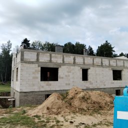 Baldyg Bud - Budowany dom jednorodzinny z białych bloczków, z widocznymi fundamentami i otworami okiennymi, pracownik na stropie, kominy, hałdy piasku na pierwszym planie.
