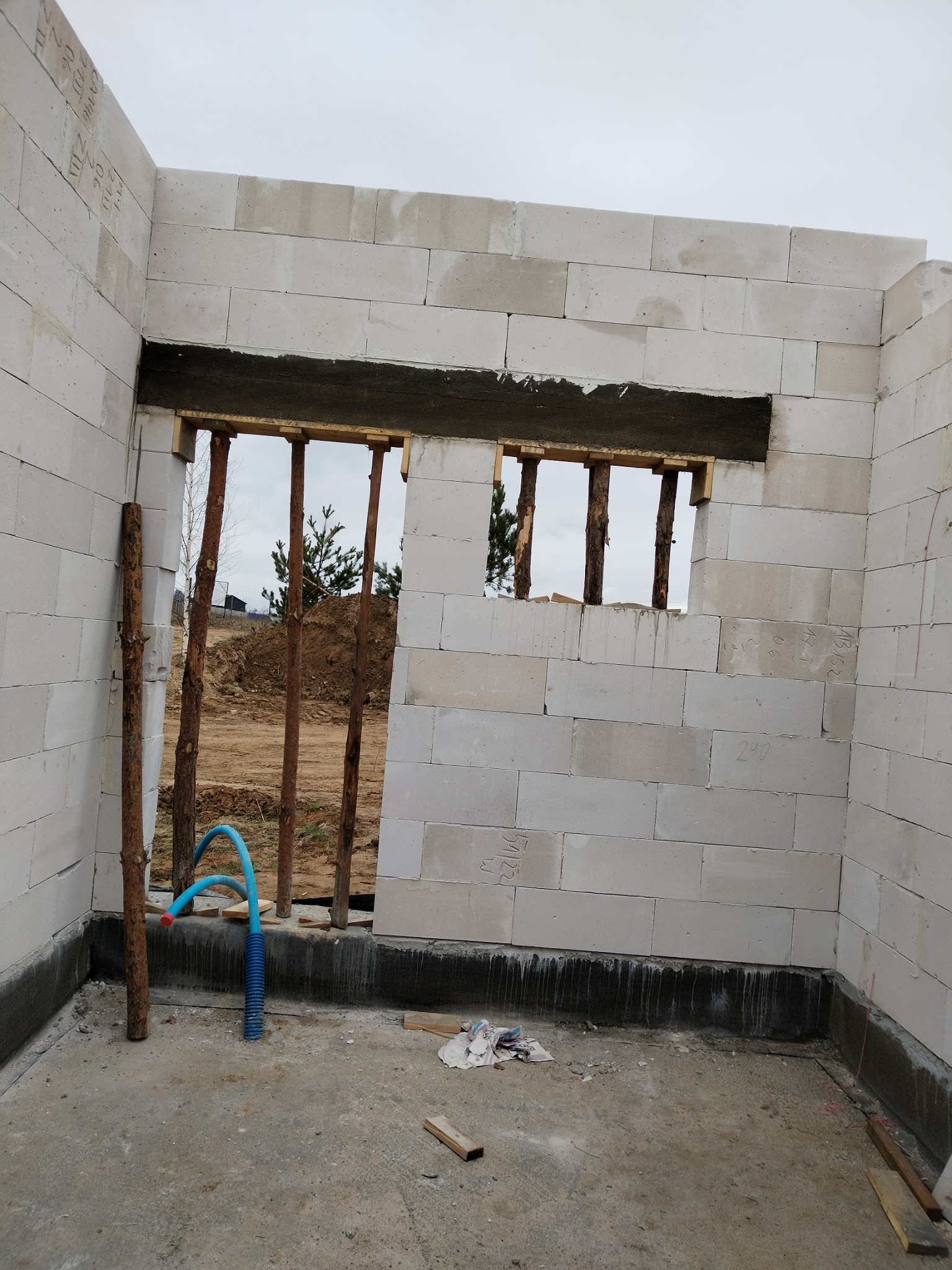 Wnętrze surowego domu z betonu komórkowego w trakcie budowy, widoczne fragmenty ścian z otworami okiennymi zabezpieczonymi drewnianymi stemplami, oraz niebieski peszel na posadzce.