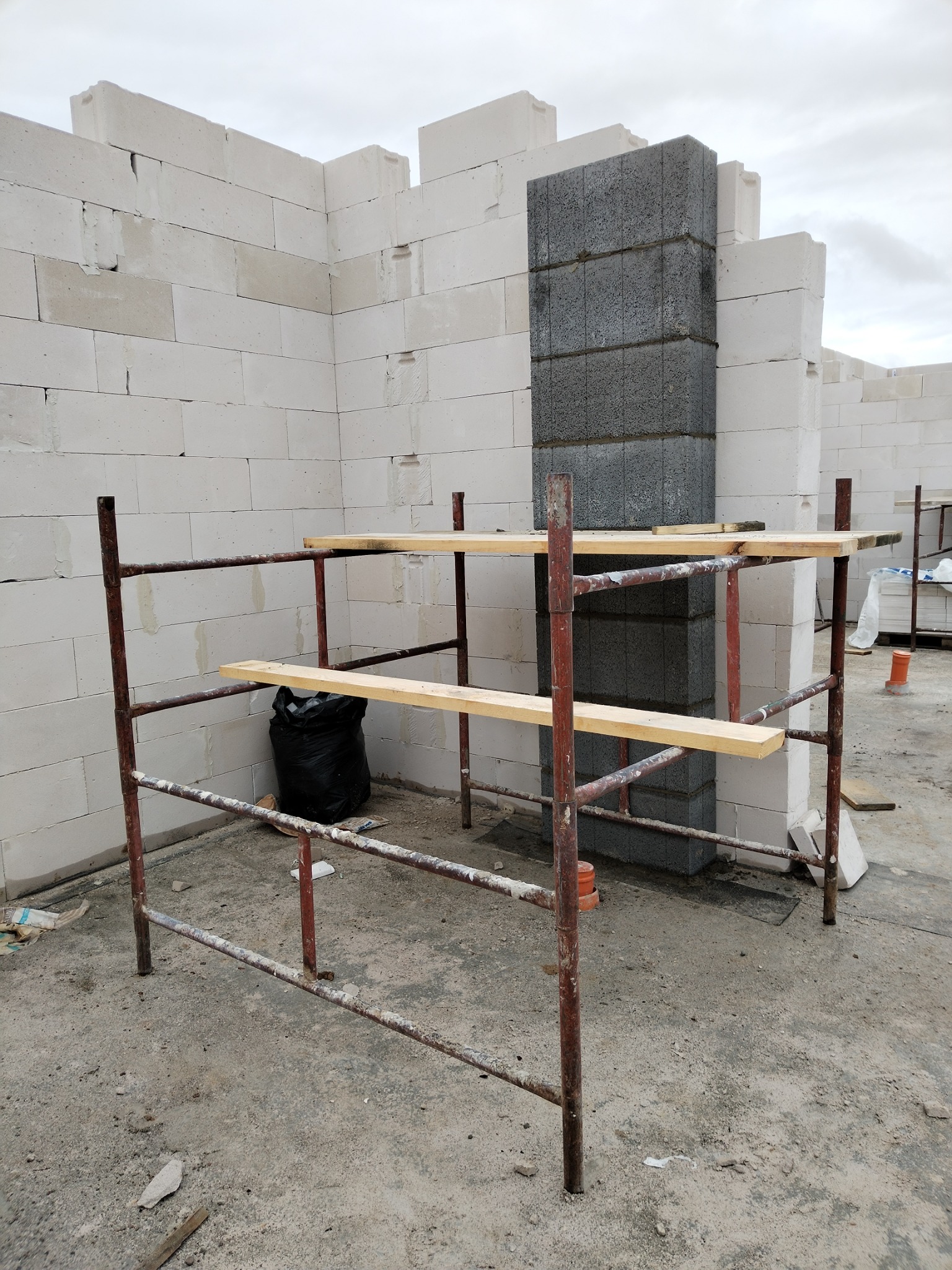 Wznoszone ściany domu z betonu komórkowego Ytong i bloczków szarych, widoczne rusztowanie z drewnianymi pomostami roboczymi, czarny worek na śmieci i fragmenty materiałów budowlanych na podłożu.