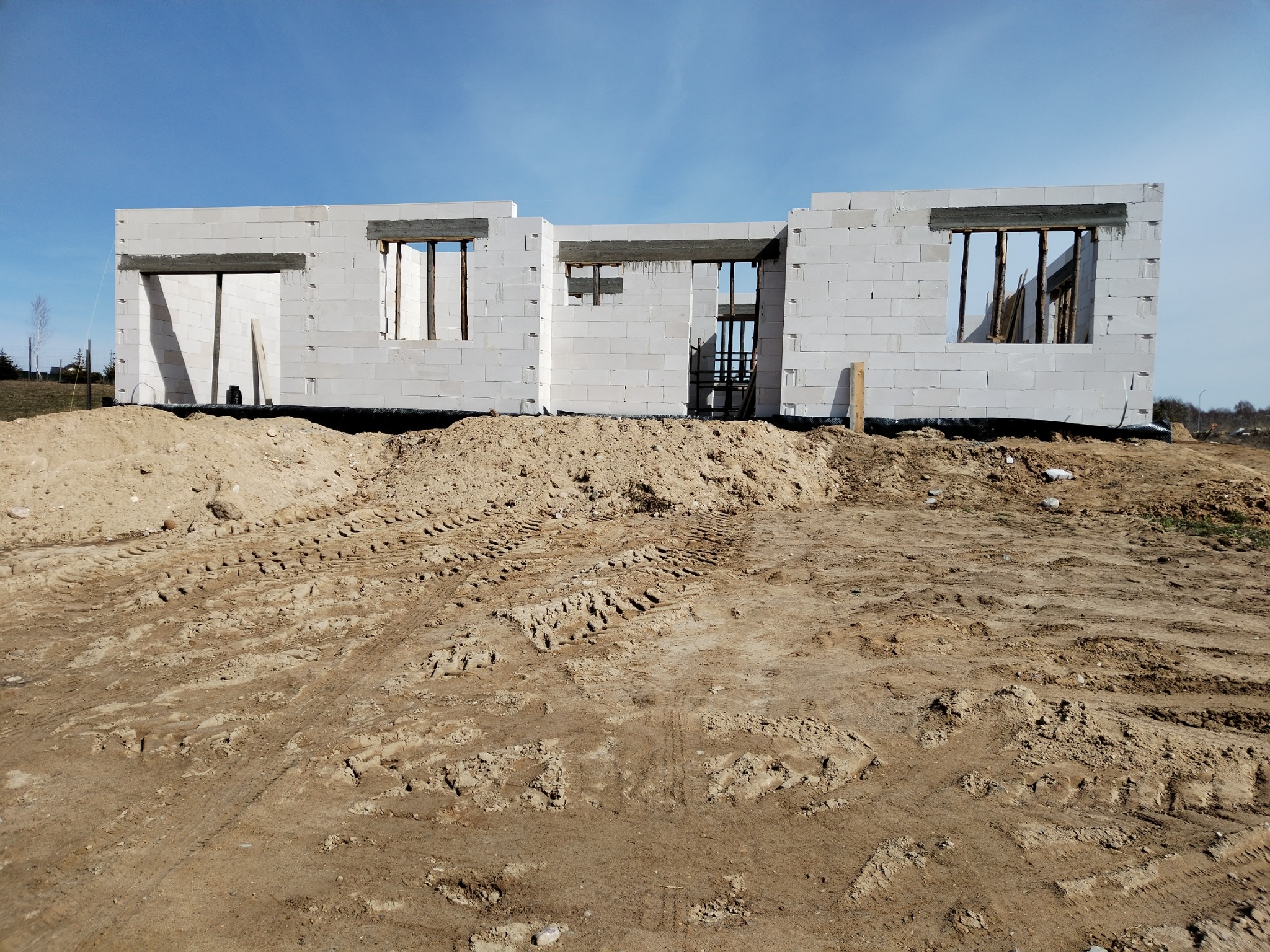 Surowy stan domu z białych bloczków, widoczne otwory okienne i drzwiowe, otoczenie z ziemi z wyraźnymi śladami opon, błękitne niebo w tle.