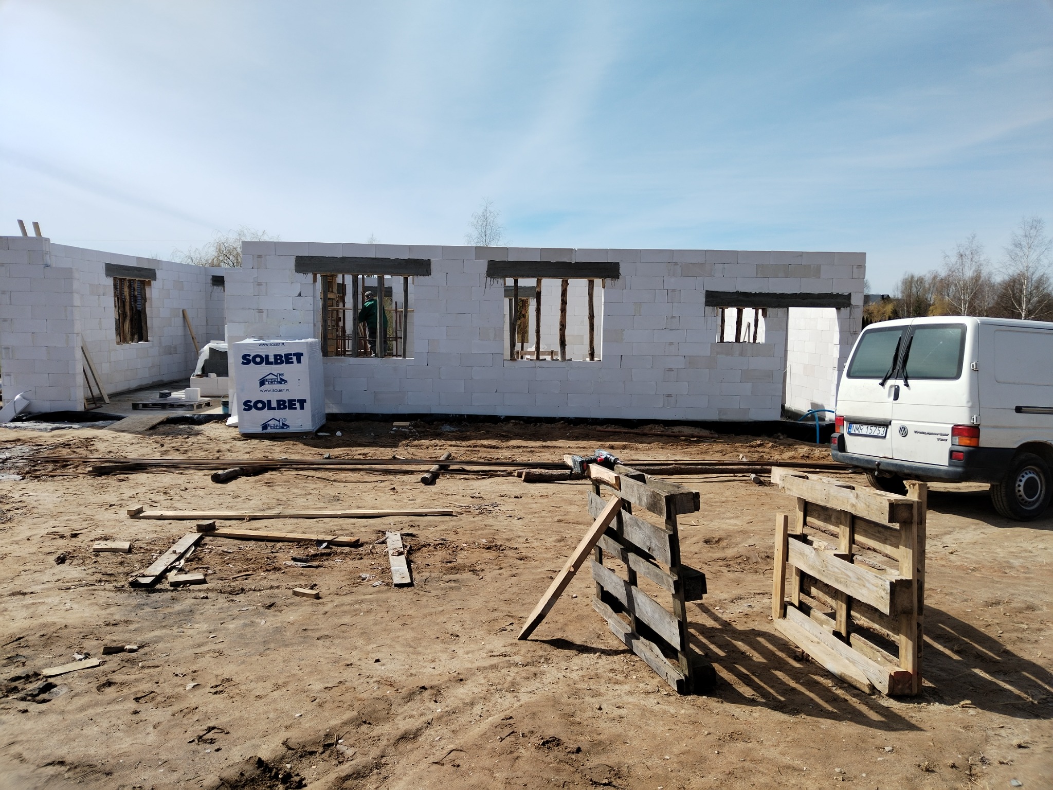 Wznoszenie ścian domu z betonu komórkowego w Sorkwitach, widoczny murarz w oknie, paleta z bloczkami Solbet i biały bus na placu budowy.