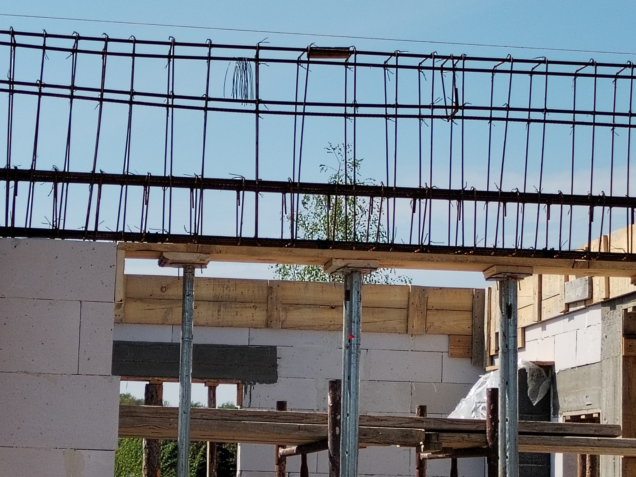 Wznoszenie ścian z betonu komórkowego, widoczne zbrojenie stropu i drewniane szalunki, w tle zieleń drzew.