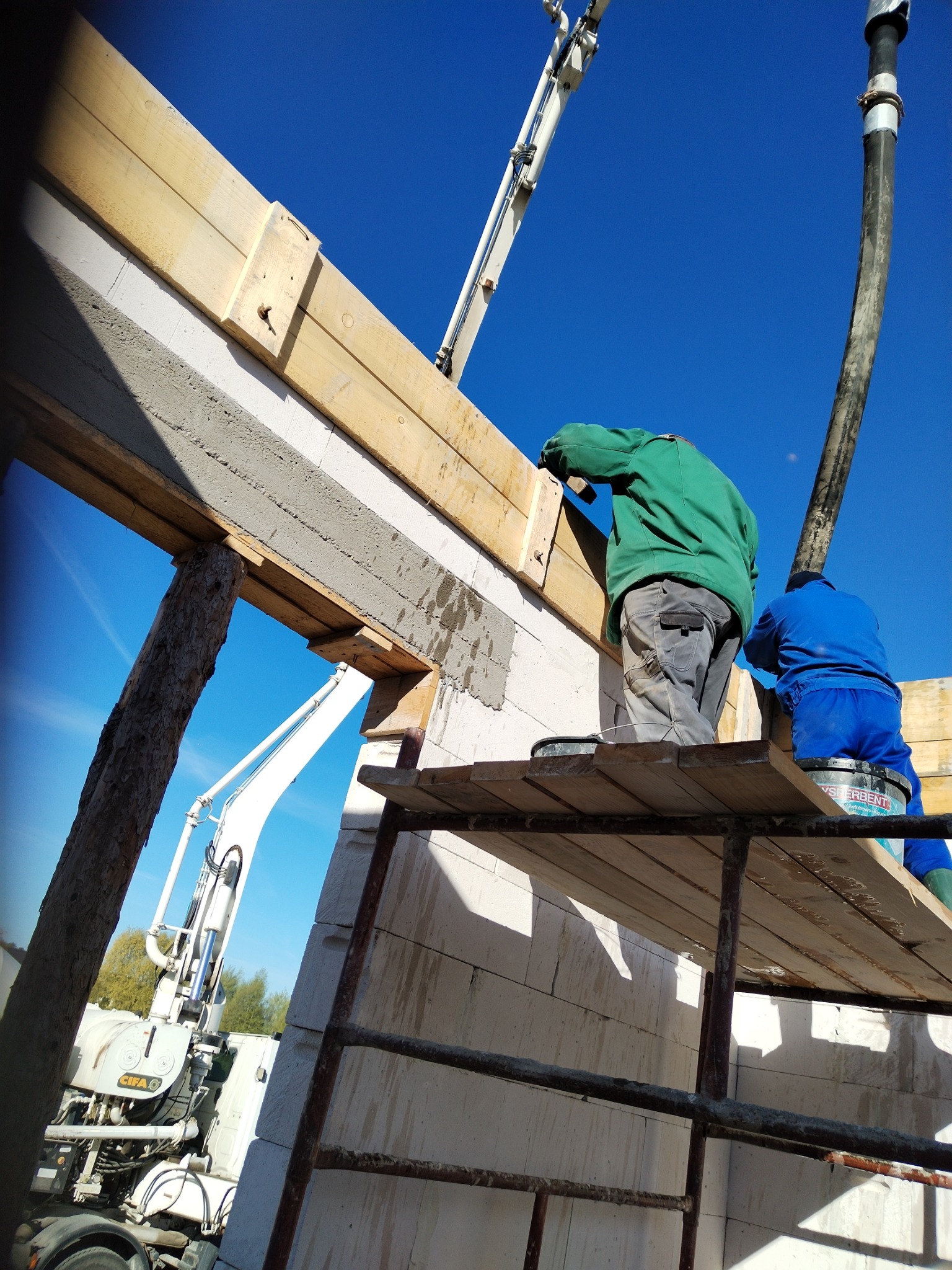 Murowanie ściany z betonu komórkowego. Widoczny fragment ściany, rusztowanie, pracownicy i maszyna do podawania betonu. Ujęcie z dołu, niebo w tle.