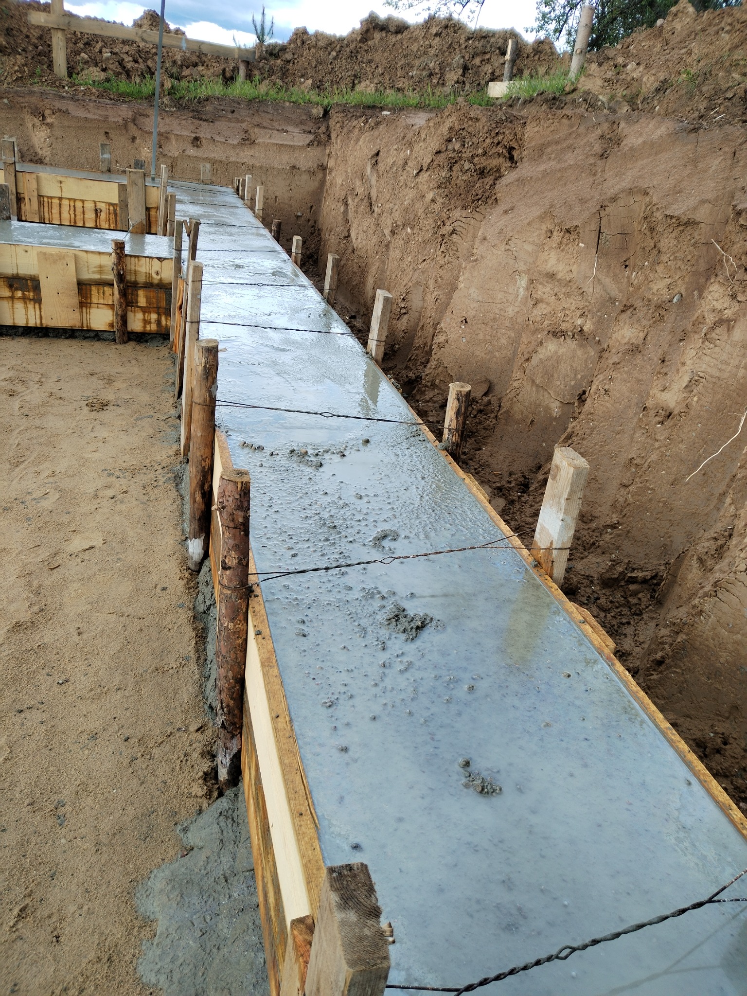 Wylany beton w szalunku drewnianym tworzący fundament. Widoczny wykop ziemny i drewniane podpory. Świeży beton z widocznymi pęcherzykami powietrza.