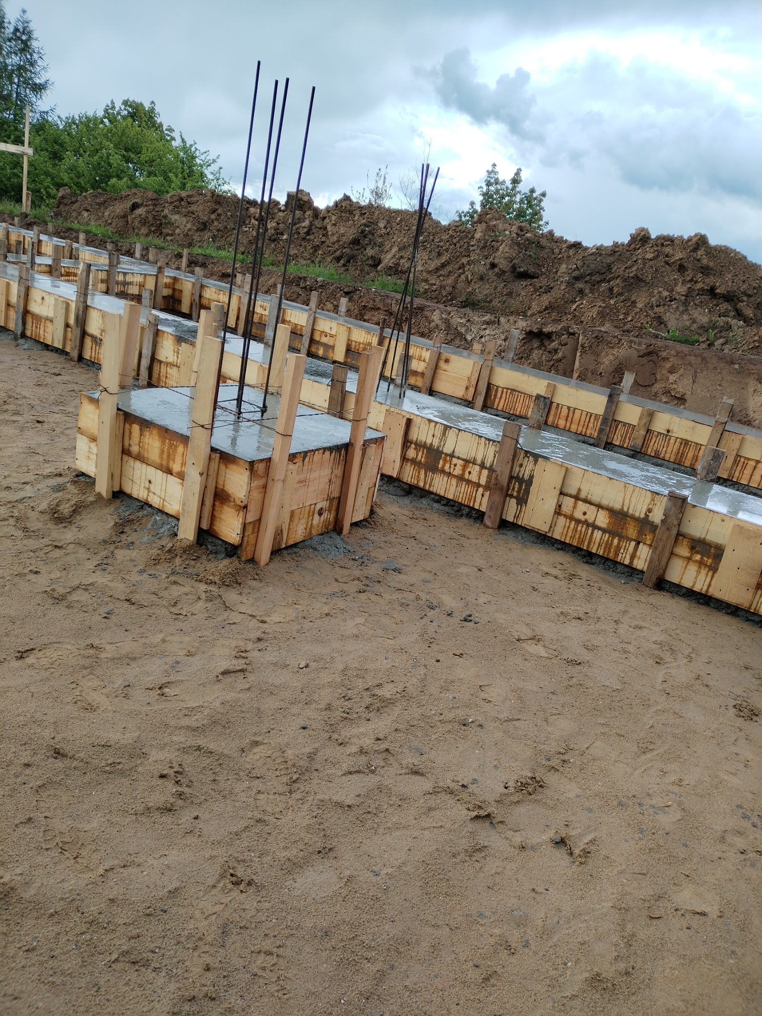 Zalane betonem szalunki fundamentowe z wystającymi prętami zbrojeniowymi, widok z góry na piasek i ziemię. Na horyzoncie zieleń drzew i pochmurne niebo.
