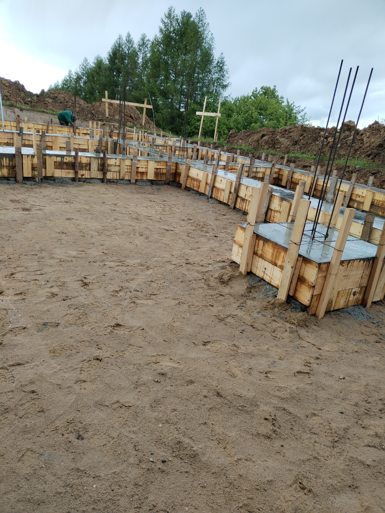 Wykonanie szalunku pod fundamenty z wystającymi prętami zbrojeniowymi na piaszczystym podłożu. W tle widoczne drzewa i kopiec ziemi.