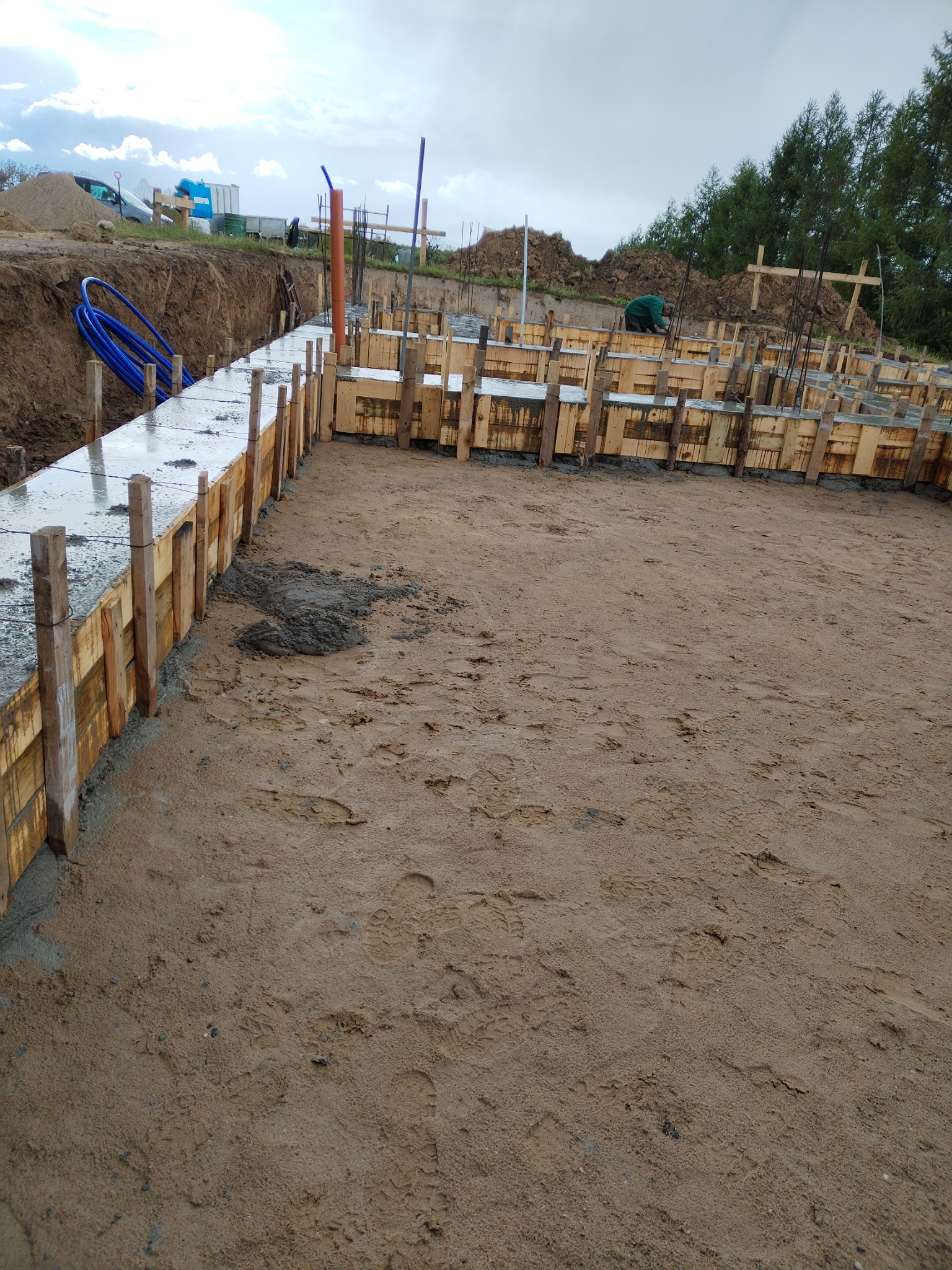 Wykop pod fundamenty z deskowaniem i zalanym betonem. Widoczne wystające pręty zbrojeniowe, rury instalacyjne i ślady stóp na piasku. Prace budowlane w toku.
