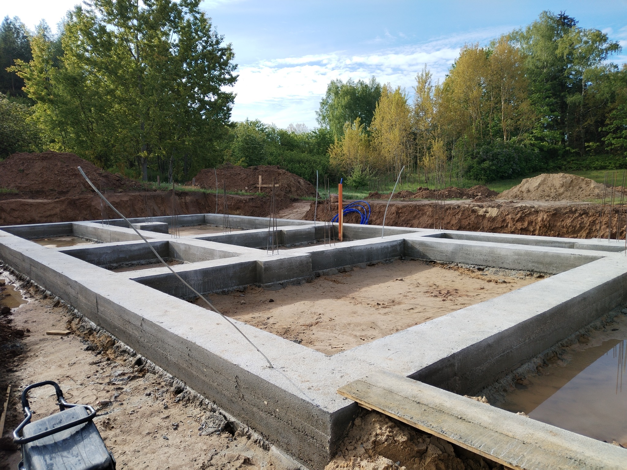 Zalane betonem fundamenty domu na działce z drzewami w tle. Widoczne zbrojenia i wykop. Na pierwszym planie wózek budowlany.