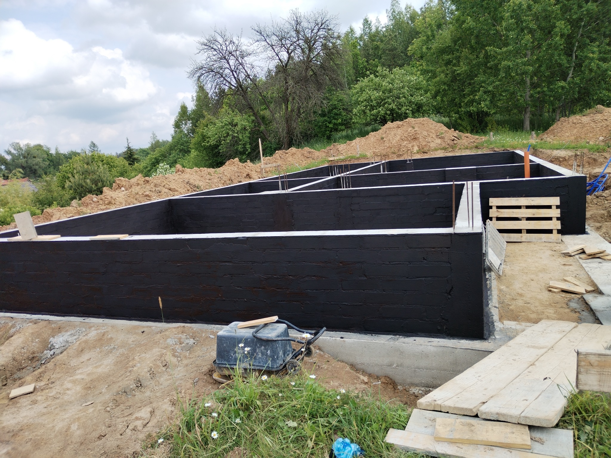 Zabezpieczone, czarne fundamenty budynku w trakcie budowy. Widoczne zbrojenia, izolacja pionowa i teren wokół fundamentów. Na pierwszym planie ubijarka.