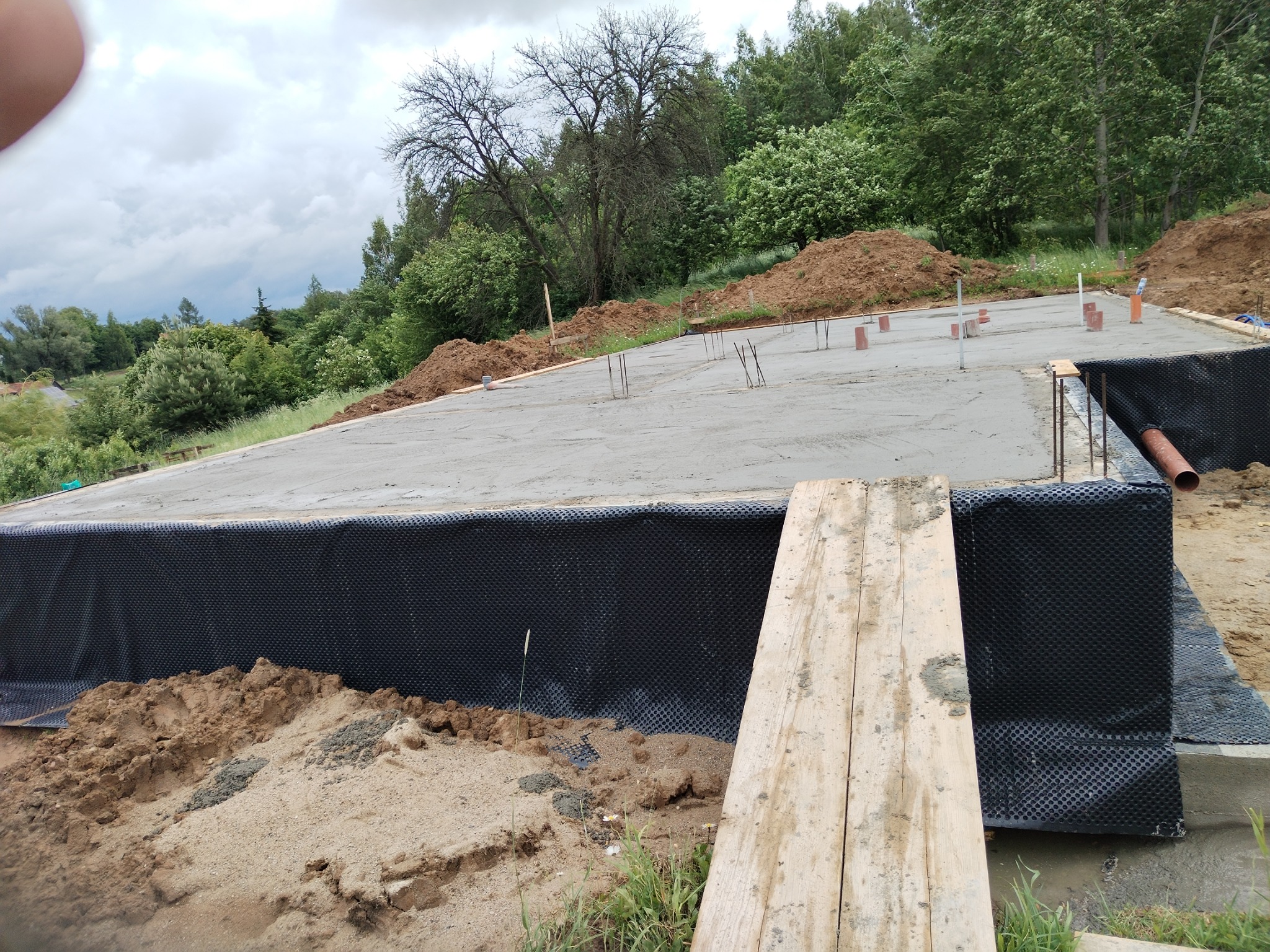 Zalany betonowy fundament z hydroizolacją w postaci czarnej folii kubełkowej, widoczne pręty zbrojeniowe i deska na pierwszym planie. Tło to zielony teren i pochmurne niebo.