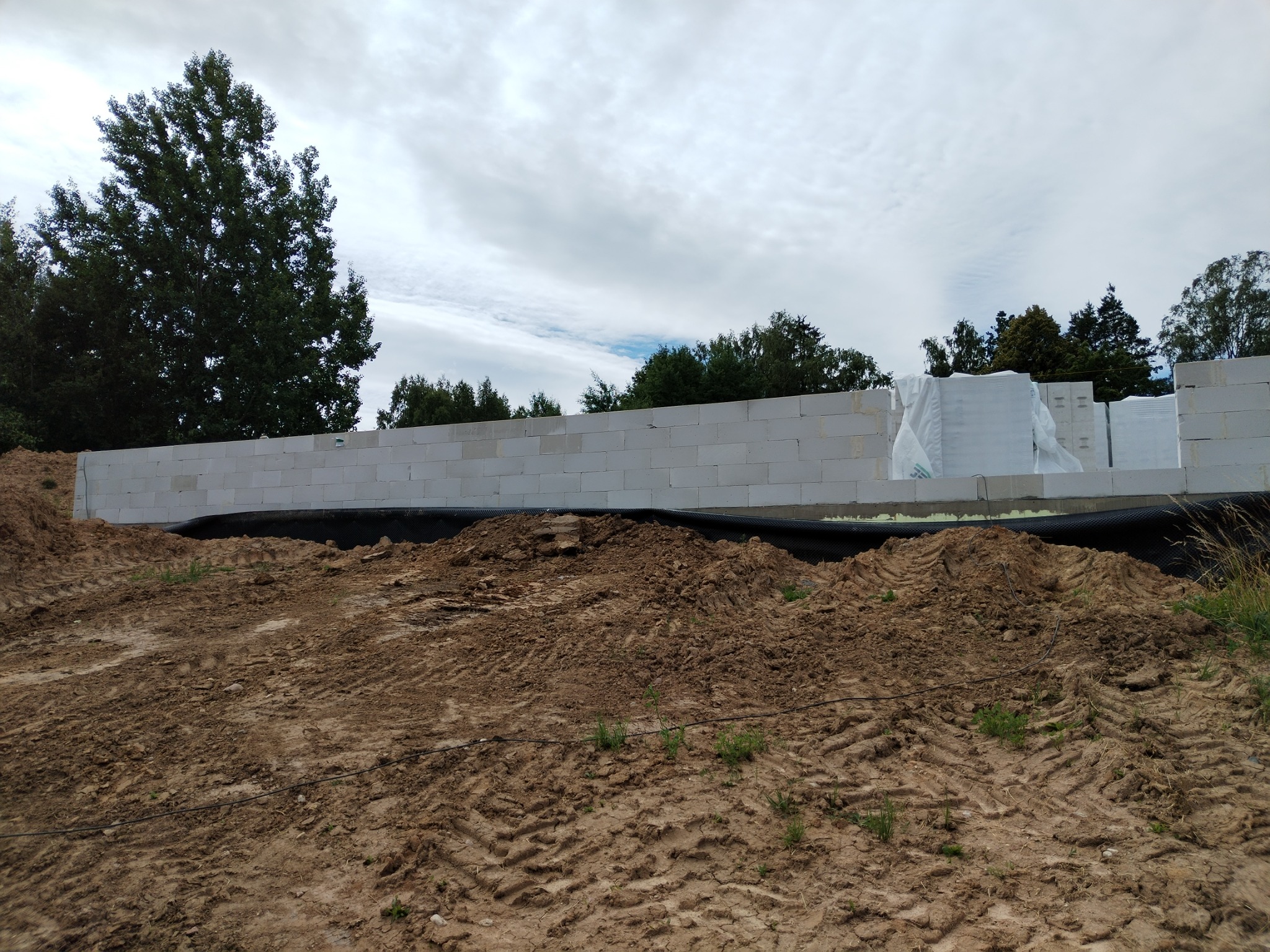 Wznoszone ściany z białych bloczków na fundamencie, zabezpieczone czarną folią. Na paletach widoczne materiały budowlane w folii ochronnej. Krajobraz z drzewami w tle.