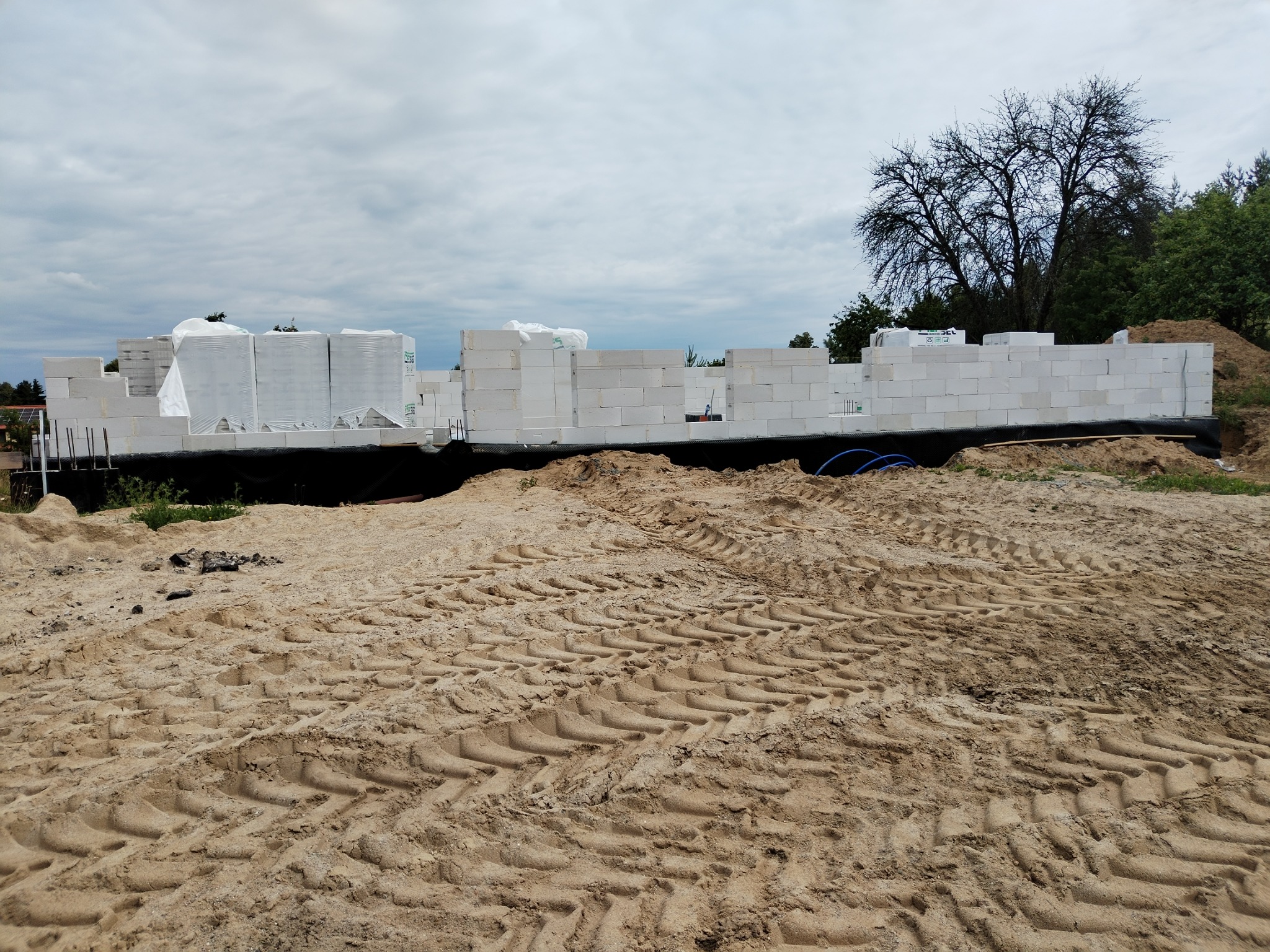 Budowa domu z białych bloczków, widoczne fundamenty i pierwsze ściany na tle piaszczystego terenu i drzew. Częściowo zapakowane bloczki czekają na użycie.