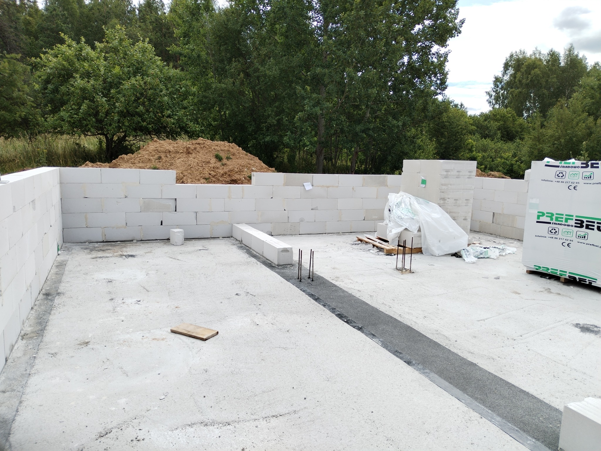 Budowa domu: ściany z bloczków betonu komórkowego Ytong na fundamencie. Widoczne zbrojenia i palety z materiałem budowlanym. W tle drzewa i usypana ziemia.