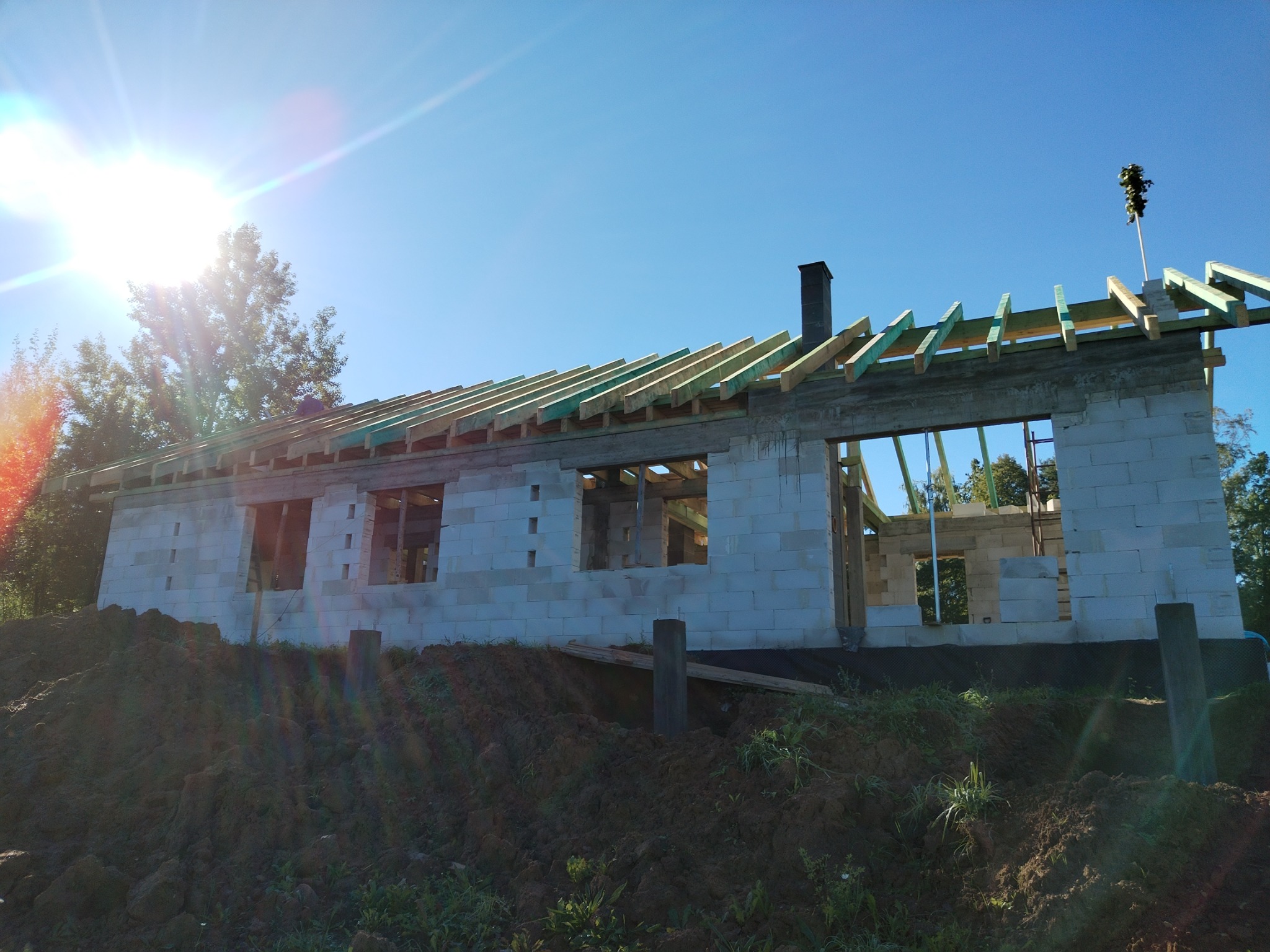 Budowa domu z bloczków: ściany z otworami okiennymi, więźba dachowa w trakcie montażu, nasyp ziemi przed budynkiem, słoneczny dzień.