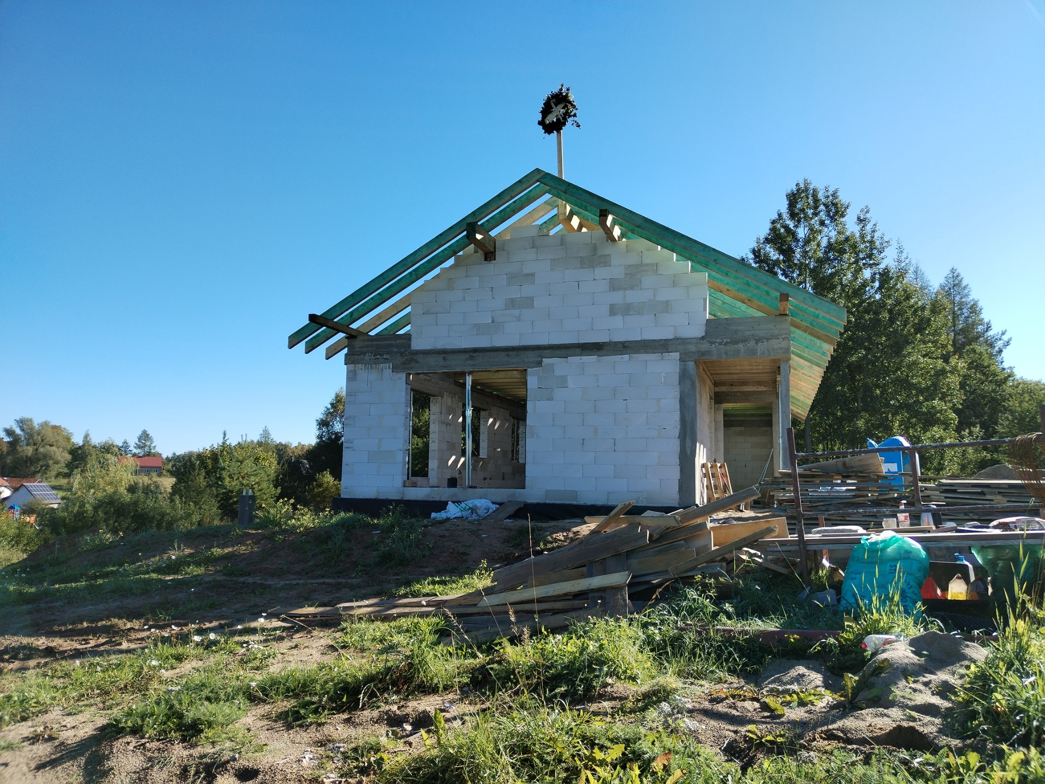 Wznoszenie domu z białych bloczków: ściany bez tynku, drewniana konstrukcja dachu pokryta zieloną folią, w tle drzewa i błękitne niebo. Wieniec na szczycie.
