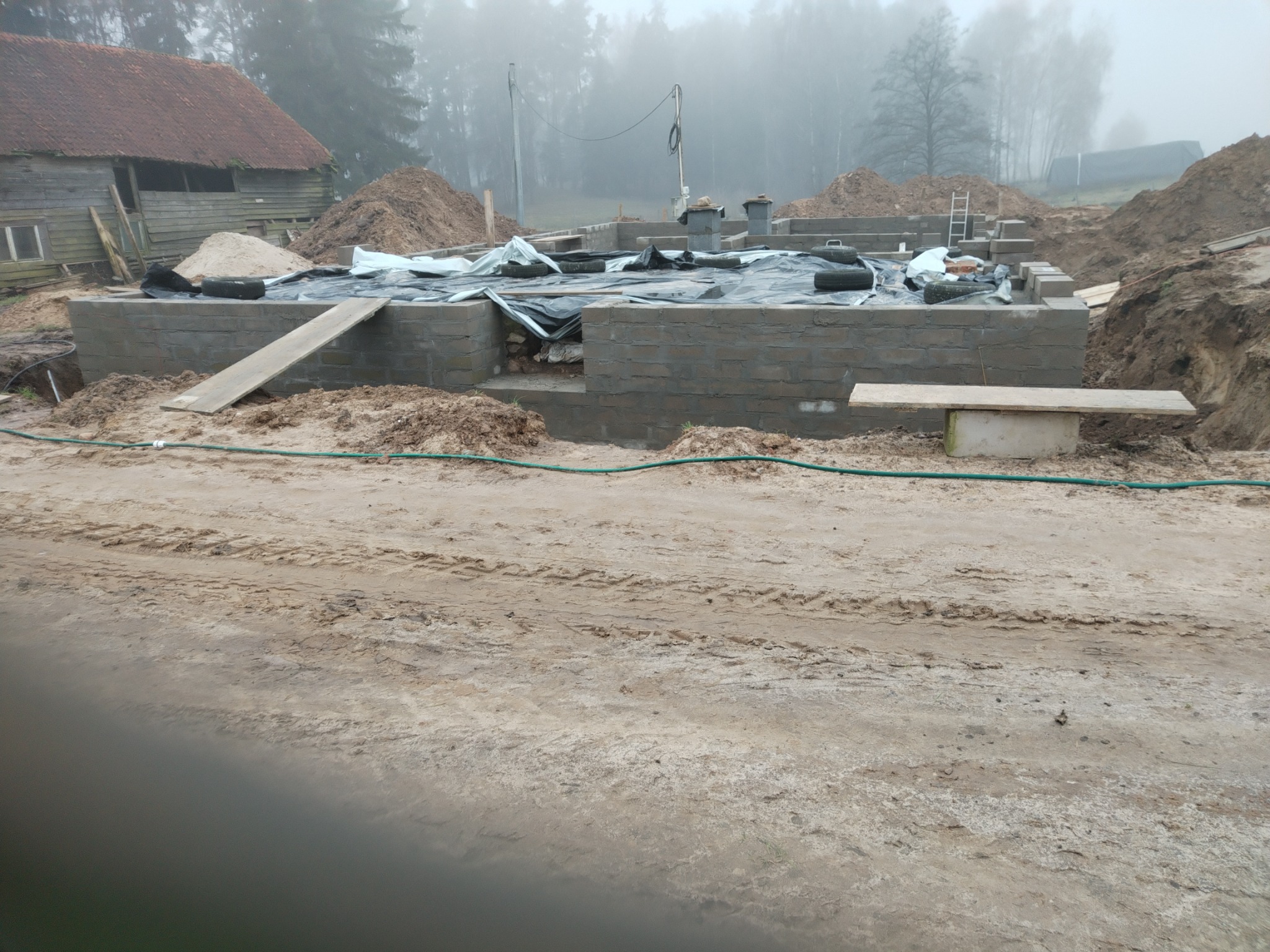 Wznoszenie fundamentów z bloczków betonowych na piaszczystym terenie, zabezpieczonych folią. W tle drewniany budynek i zalesiony teren w porannej mgle.