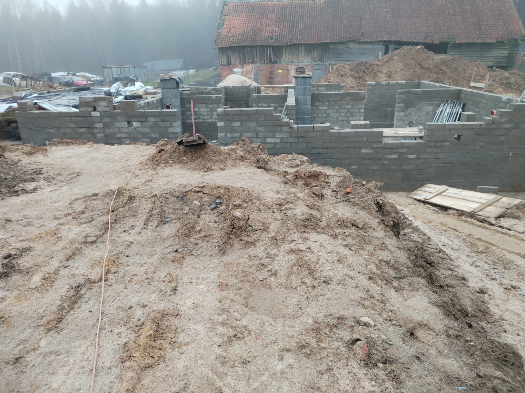 Wznoszenie ścian fundamentowych z bloczków betonowych na placu budowy. Widoczne zbrojenia i elementy wentylacyjne. W tle stary budynek gospodarczy i mgła.