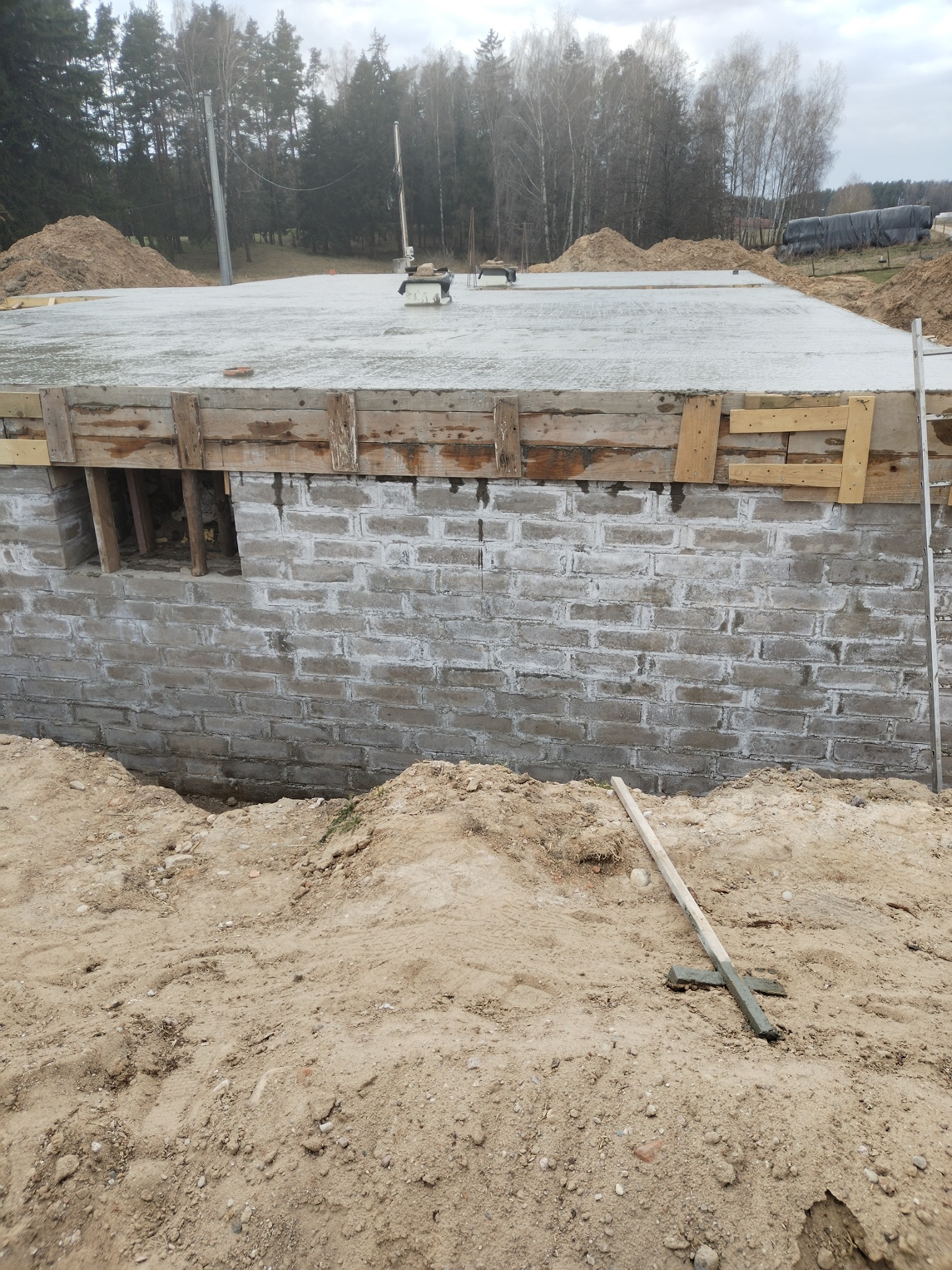 Wylany fundament z izolacją pionową i szalunkiem drewnianym. Widoczne wystające rury instalacyjne i teren budowy. W tle las.