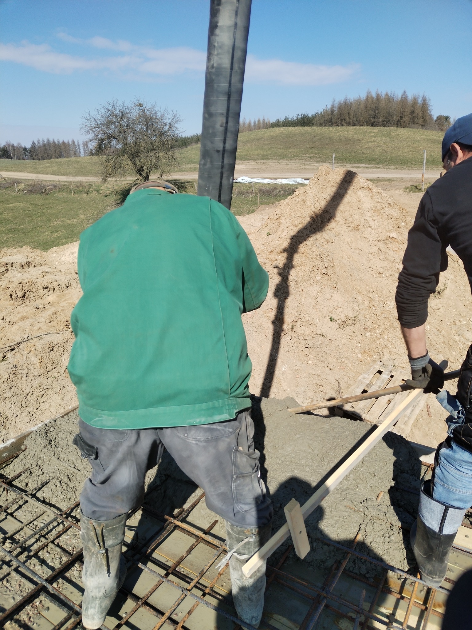 Wylewanie betonu na zbrojenie fundamentów. Widoczni dwaj pracownicy w roboczych ubraniach i kaloszach. W tle ziemia i drzewa. Słoneczny dzień.