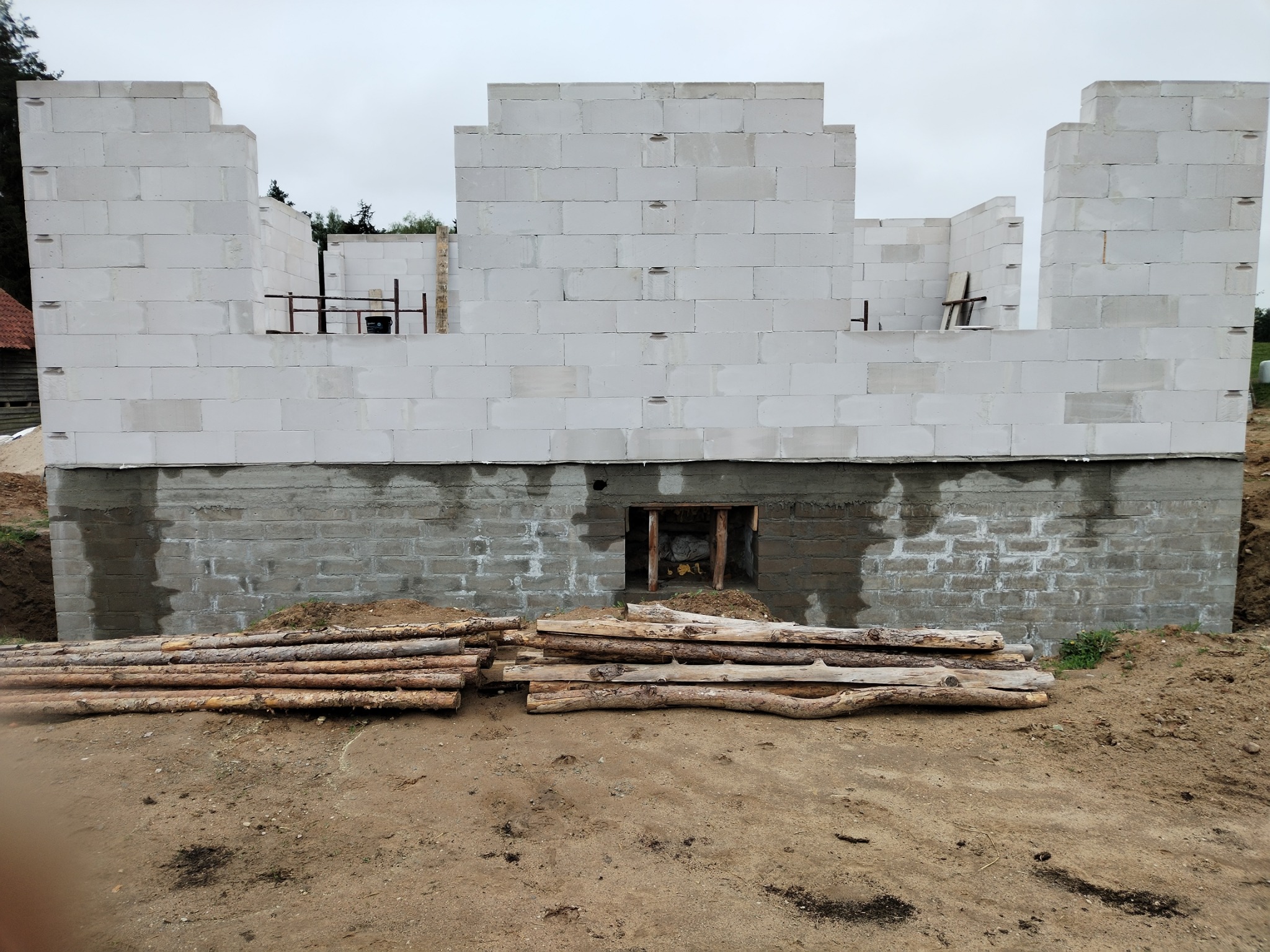 Wznoszenie ścian domu z betonu komórkowego na fundamencie. Widoczne drewniane stemple i stos drewna na ziemi. Surowy stan budowy, pochmurne niebo w tle.