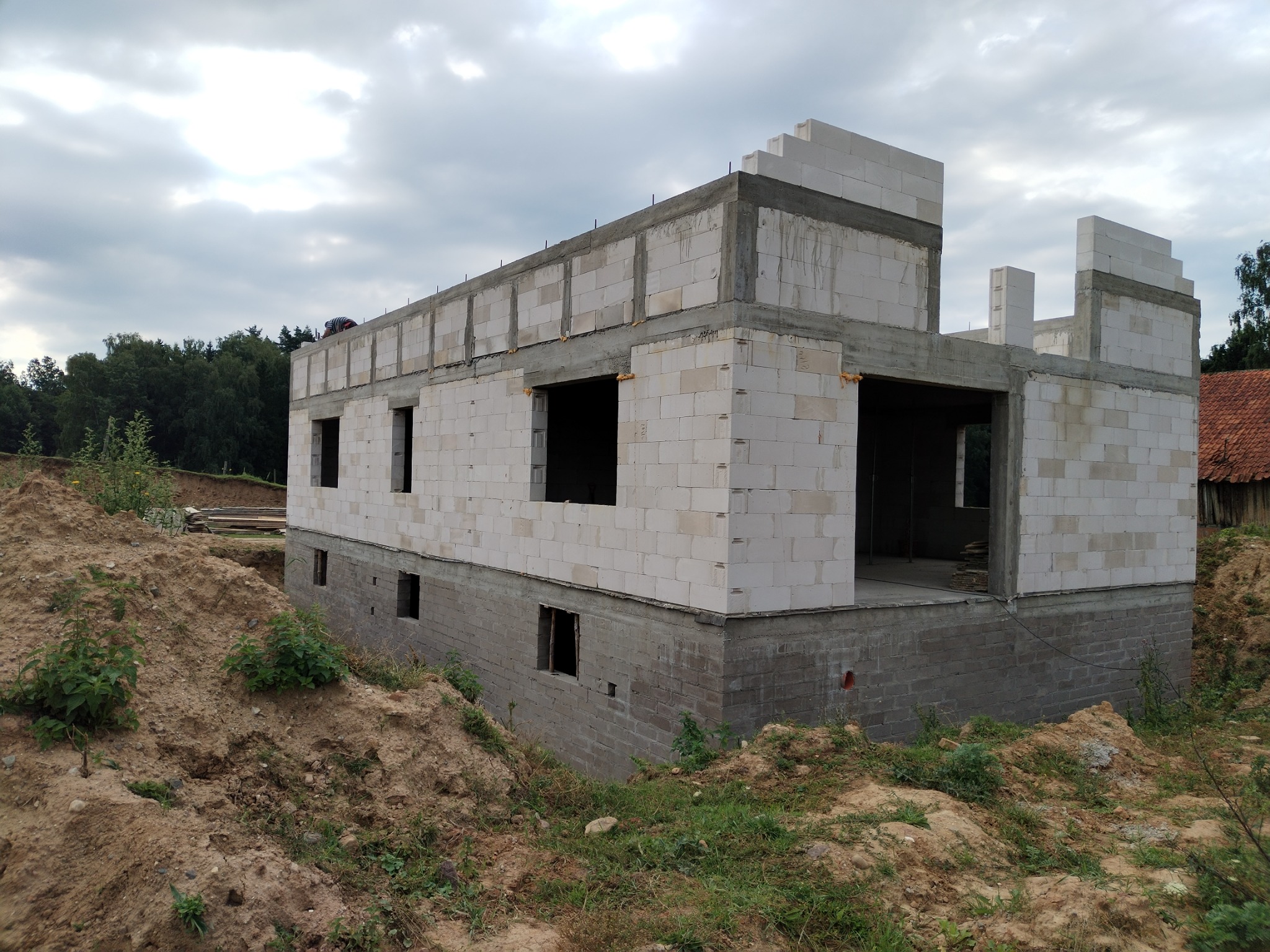 Budynek mieszkalny w trakcie budowy: parter z białych bloczków, szare fundamenty, widoczny wieniec betonowy. Rozpoczęte ściany piętra, wokół ziemia budowlana.
