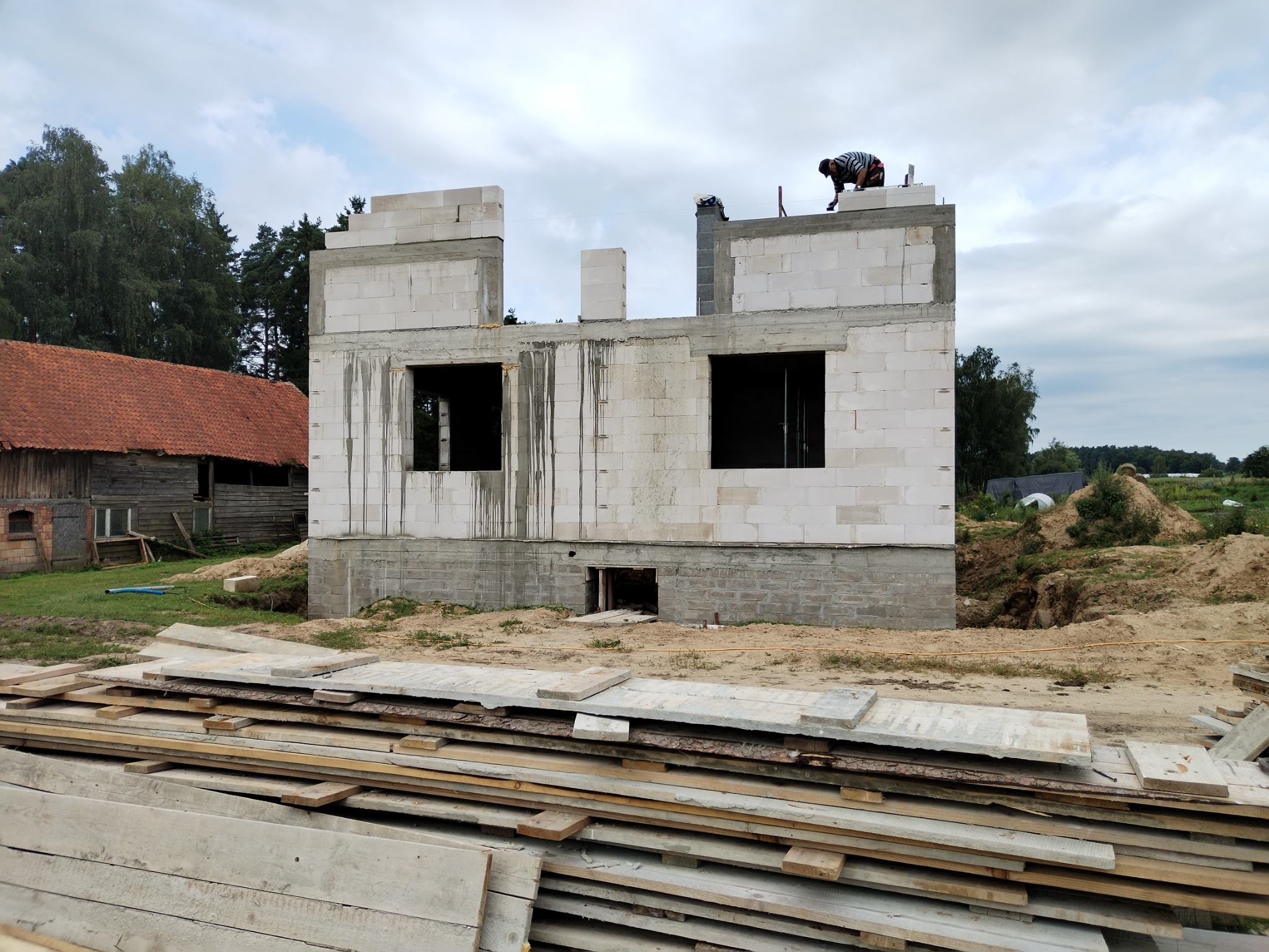 Wznoszenie ścian parteru i piętra domu z białych bloczków, z widocznym cokołem. Pracownik na górze, stos desek na pierwszym planie, w tle stary budynek z dachówką.